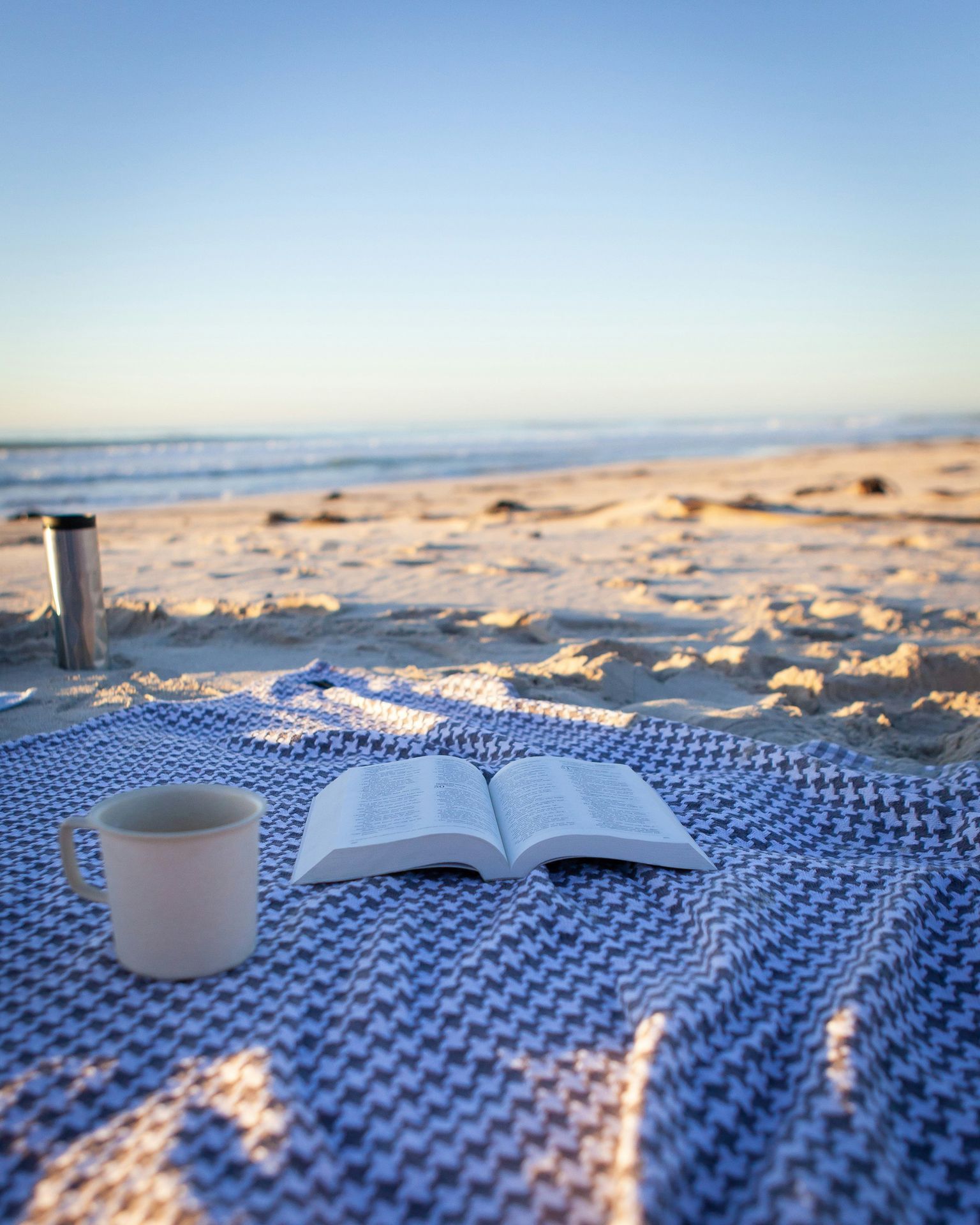 beach blanket on the a white sand beach, with an open book, and mug
