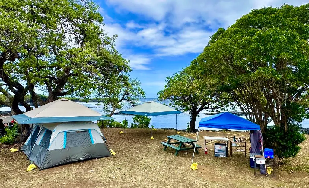 daytime fully setup campsite with tents, canopies, picnic table, kitchen area, green trees, ocean in the background, cooler, tables